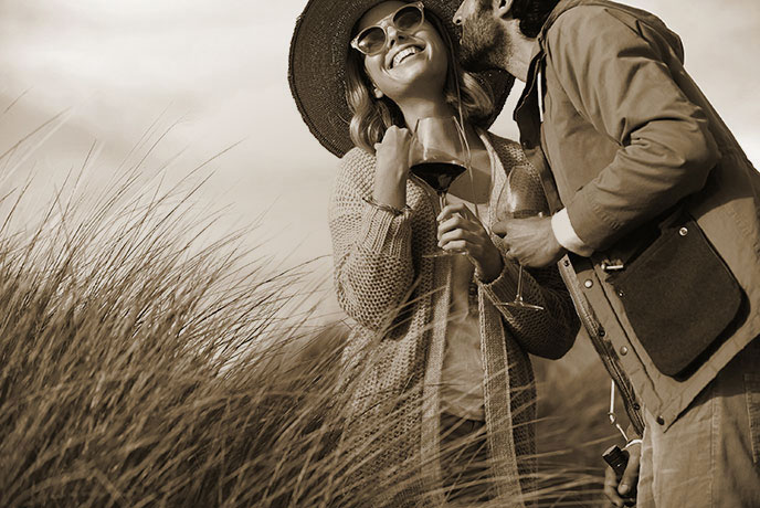 Belle Glos lifestyle sepia photo - a couple embracing while sharing a glass of wine on a wind-swept beach